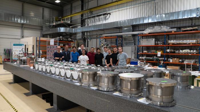 group of people standing behind a table of many cylindrical pieces of equipment that are the shape of sewing bobbins